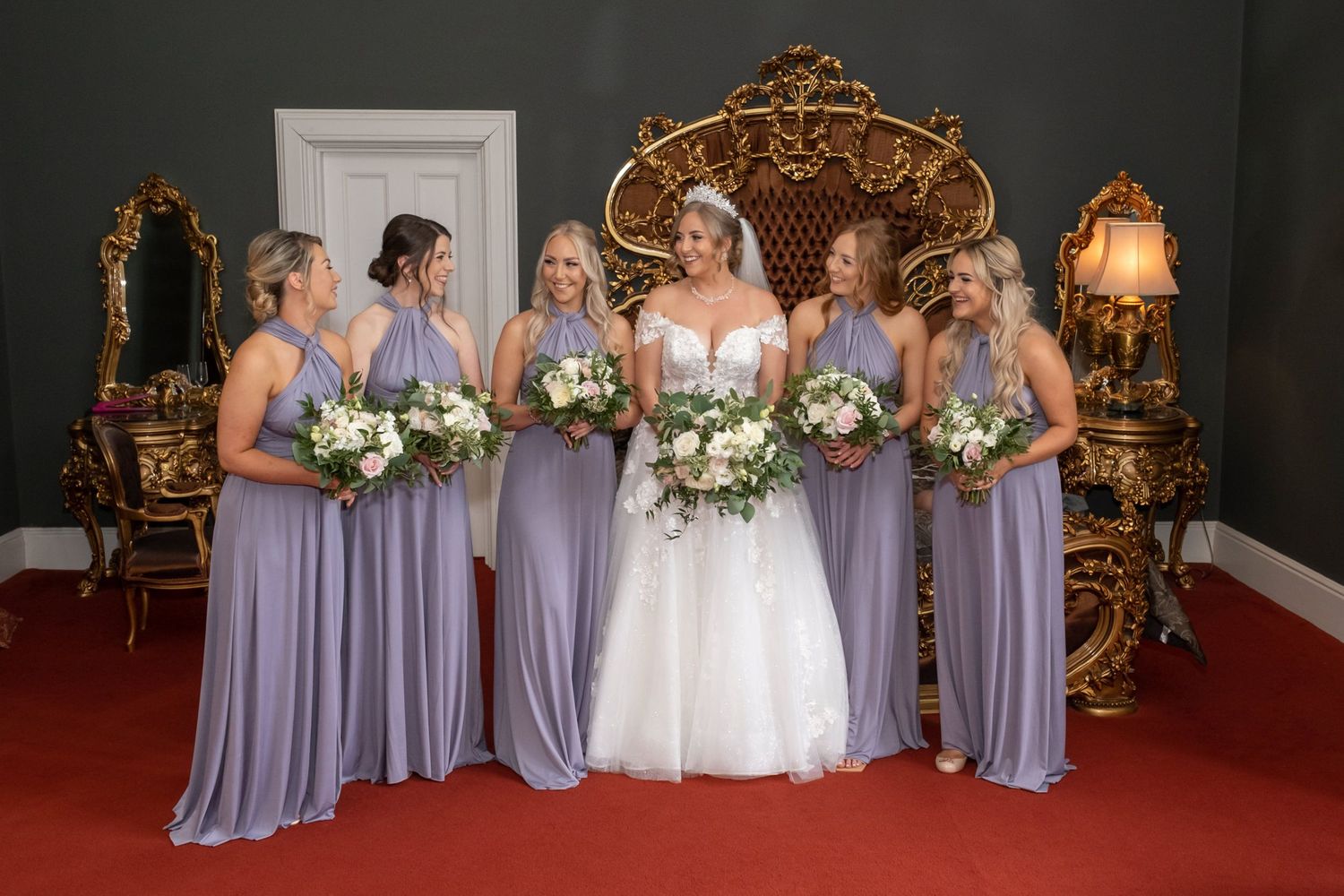 Bridesmaids in lavender dresses stand with bride in white gown against ornate gold decor at elegant venue.