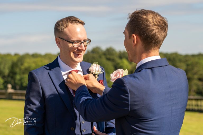 Groomsman helping adjust boutonniere on another's navy suit lapel.