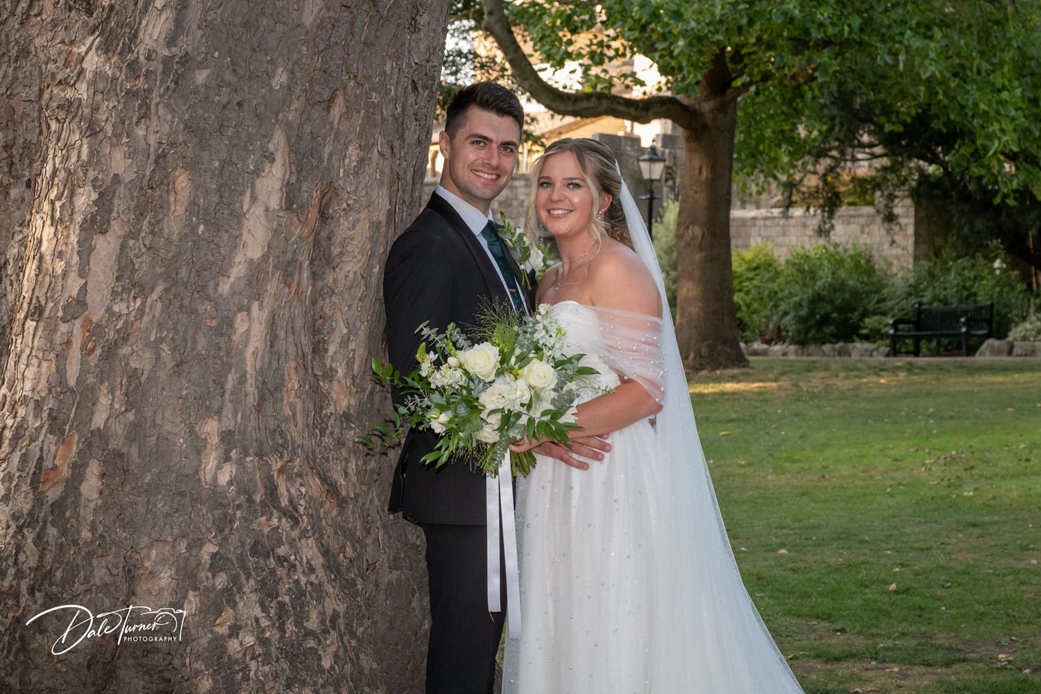 Wedding couple poses by large tree trunk in garden setting with flowing white gown.