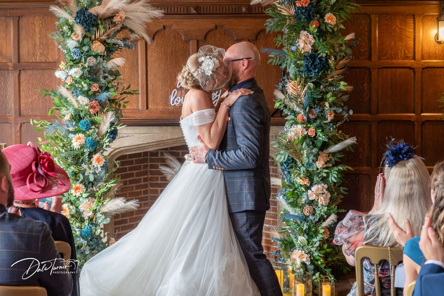Bride and groom share their first kiss at a floral arch ceremony in an elegant wood-paneled venue.