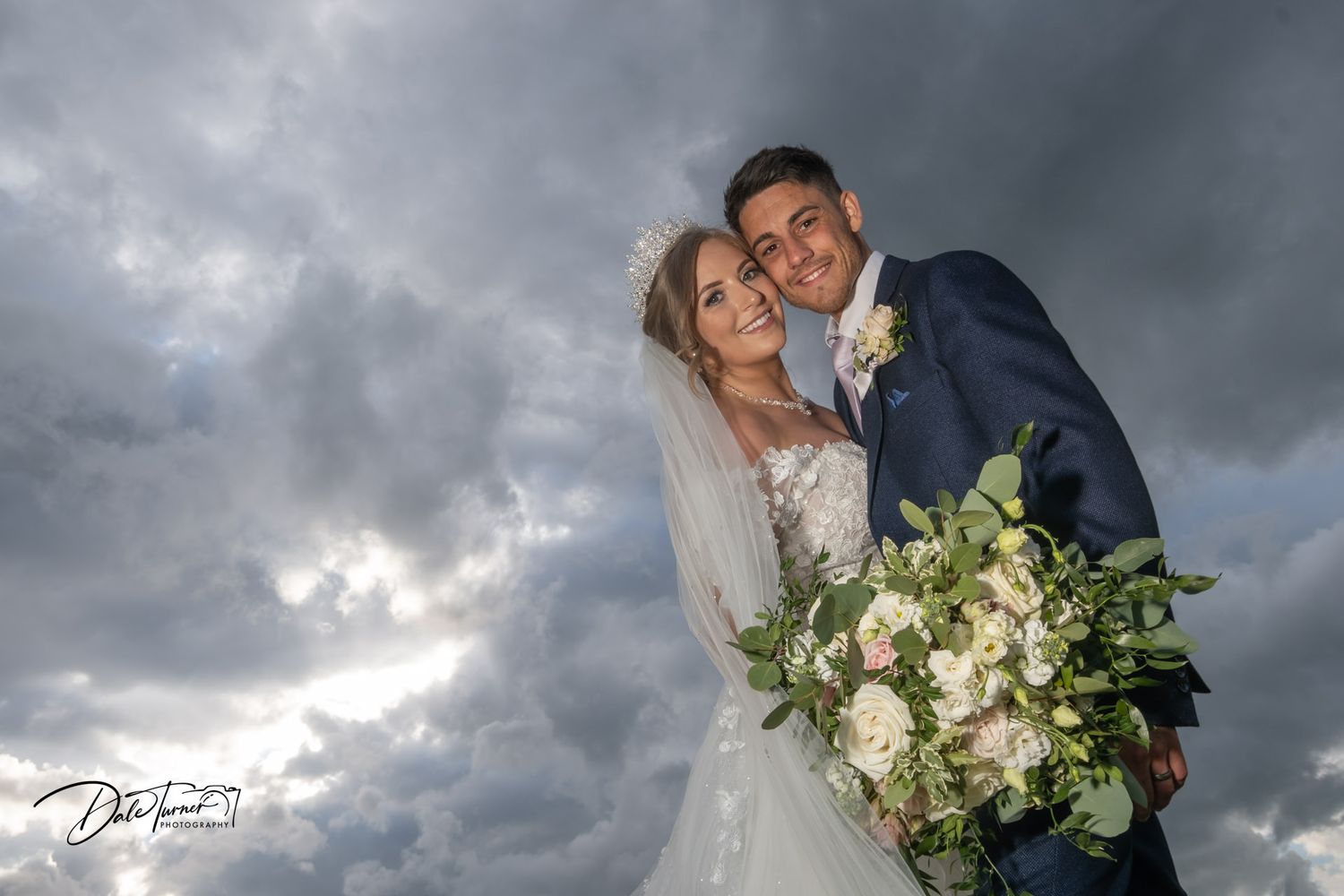 Wedding couple embracing under dramatic stormy skies with white floral bouquet.