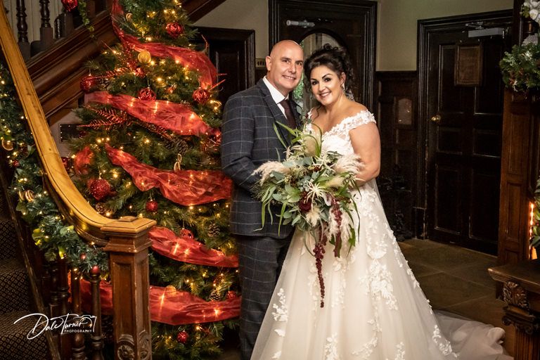 Wedding couple poses by illuminated Christmas tree on elegant wooden staircase.