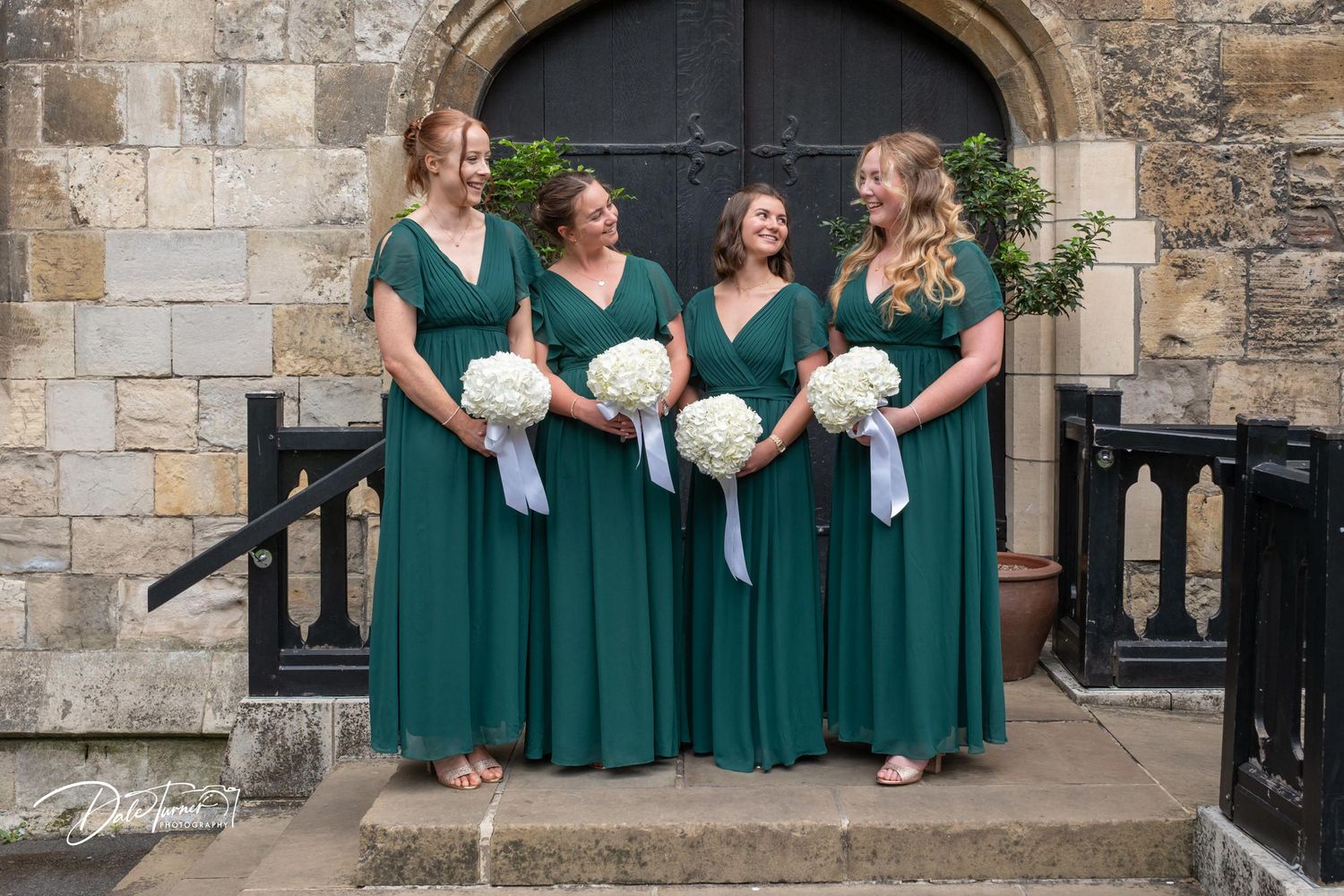 Bridesmaids in emerald green dresses pose together on stone steps at a wedding venue.