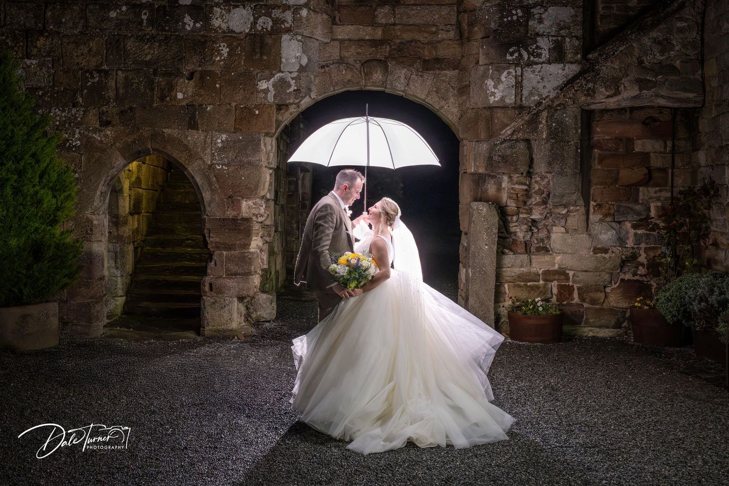 Bride and groom pose under white umbrella against rustic stone wall backdrop during wedding photoshoot.
