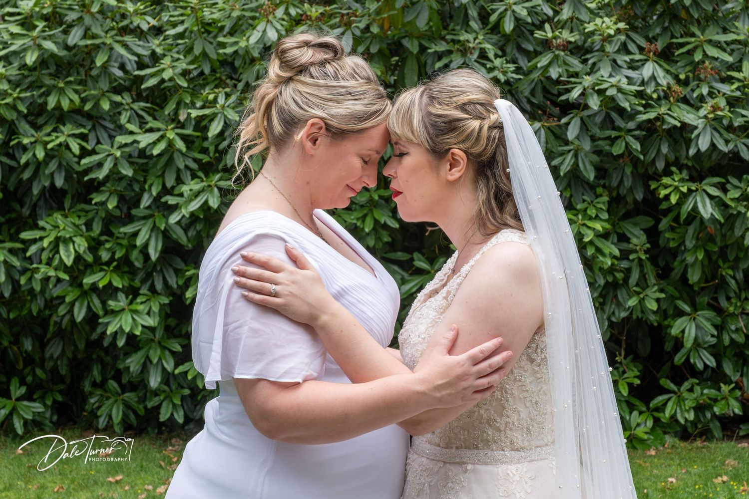 Wedding celebration group embraces outdoors against lush green foliage backdrop.