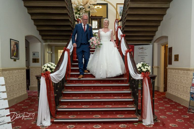 Wedding couple stands on elegant red-carpeted stairs with white fabric decorations.