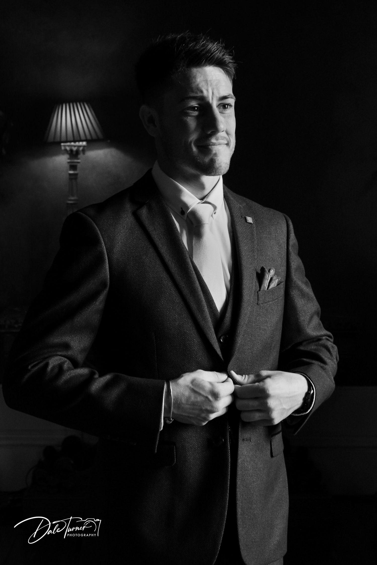 Dramatic black and white portrait of groom adjusting suit in low key lighting.