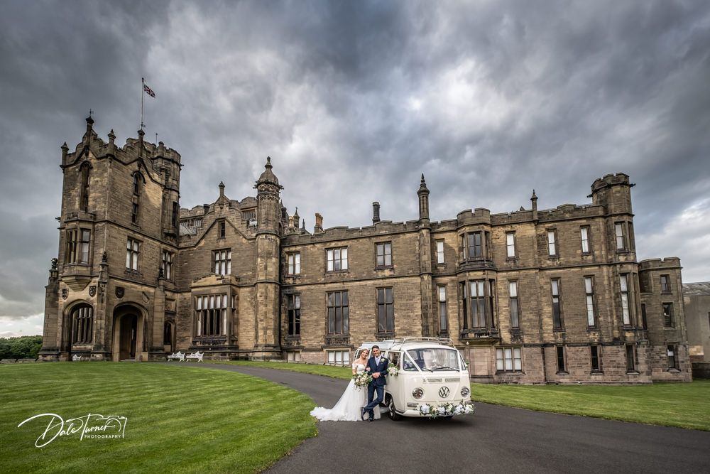 Gothic castle architecture with classic vintage car parked outside under dramatic cloudy skies.