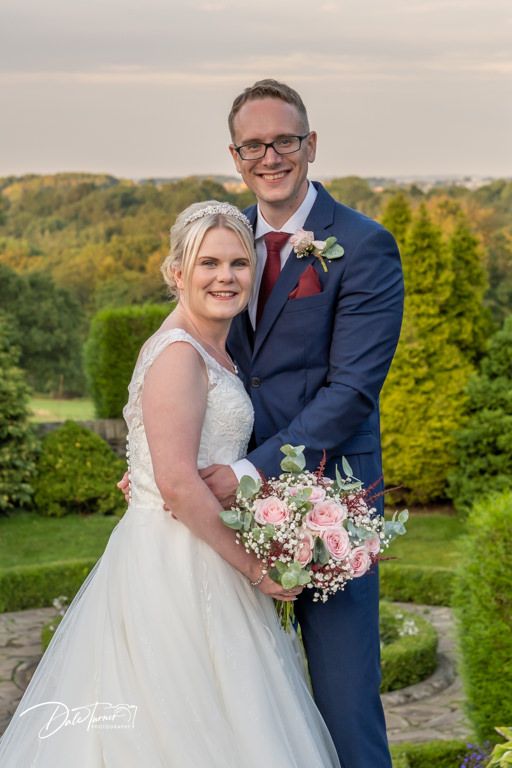 Joyful wedding couple poses with pink and white bouquet in garden setting.
