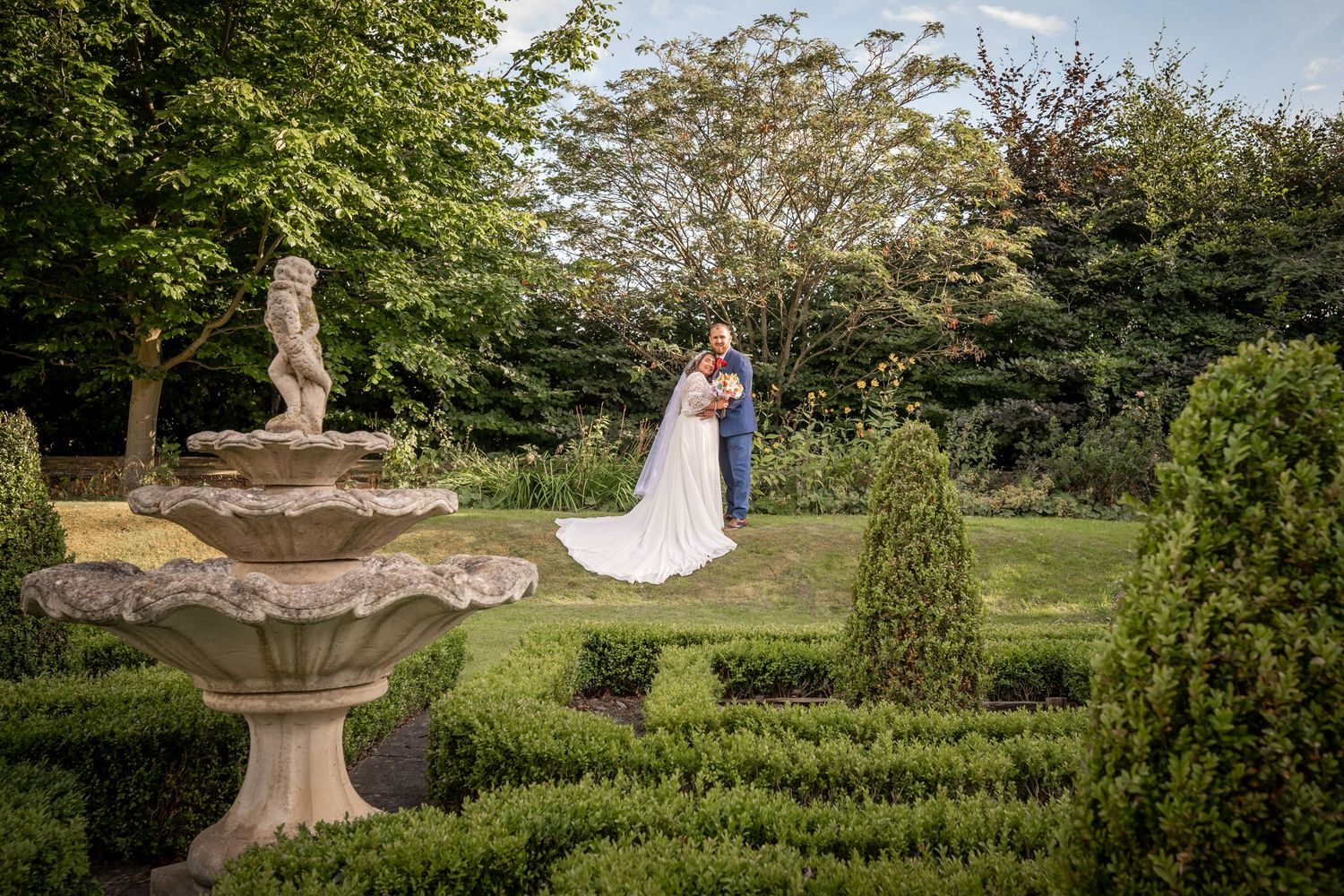 Wedding photography in a beautiful garden setting with ornate stone fountain and manicured hedges.