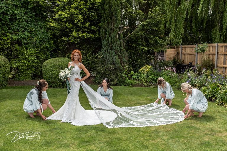 Wedding party arranges bride's long lace train for photos in garden setting.