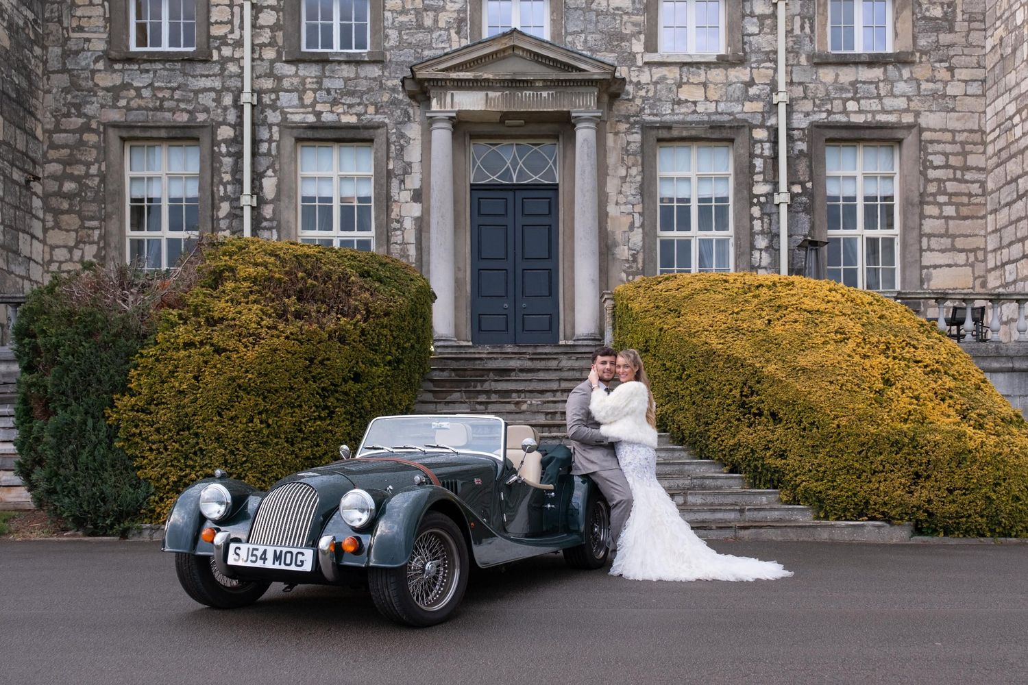 Panoramic view of a grand stone manor house with classic architecture and vintage automobiles.
