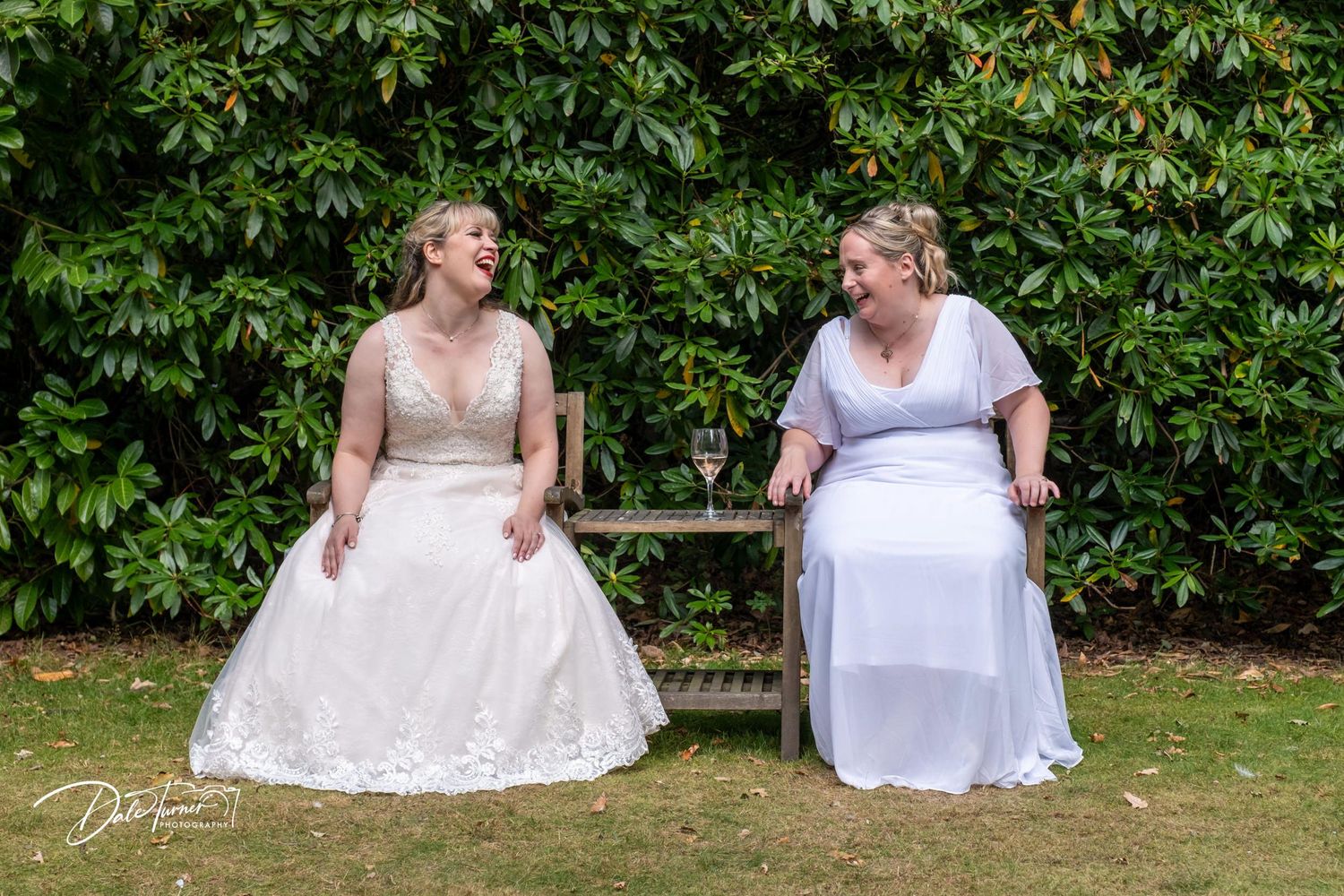 Two people in white wedding dresses sitting together on a garden bench surrounded by greenery.