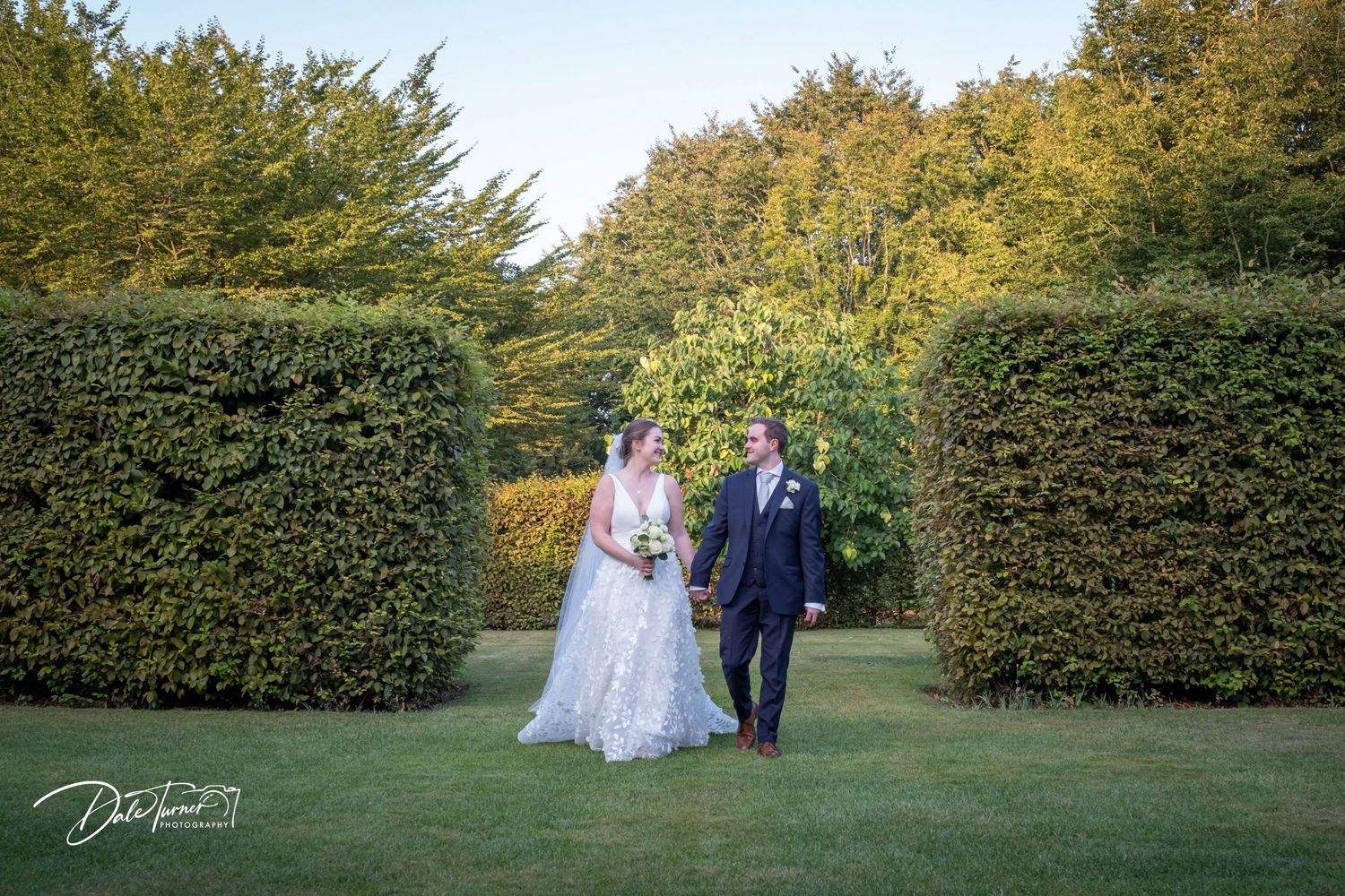 Wide landscape view of newlyweds against manicured hedges and autumn trees.