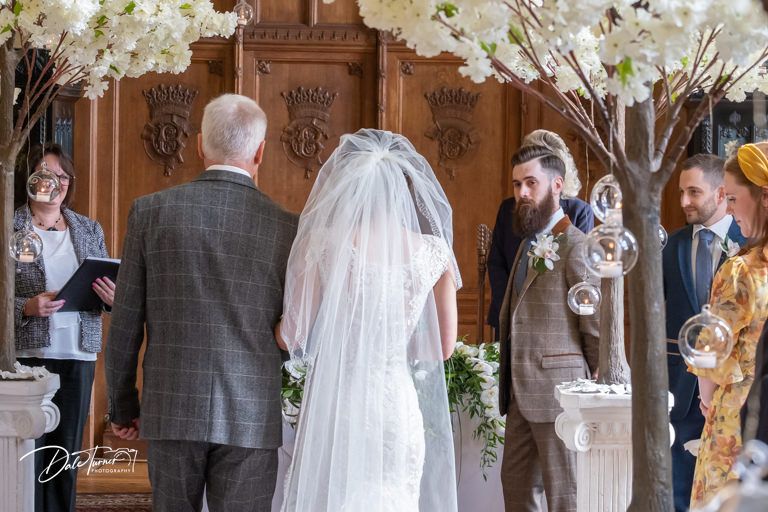 Wedding ceremony in elegant wood-paneled hall decorated with white floral arrangements.