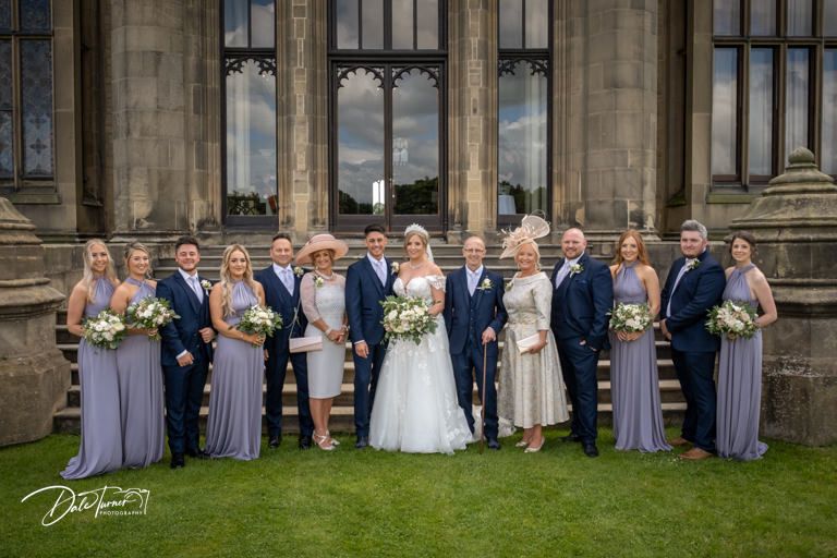 Formal wedding party lineup against historic stone building backdrop with manicured grounds.