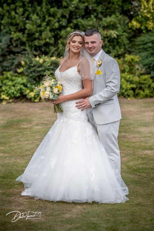 Bride in flowing white gown holding yellow bouquet poses with groom in light gray suit on garden lawn.