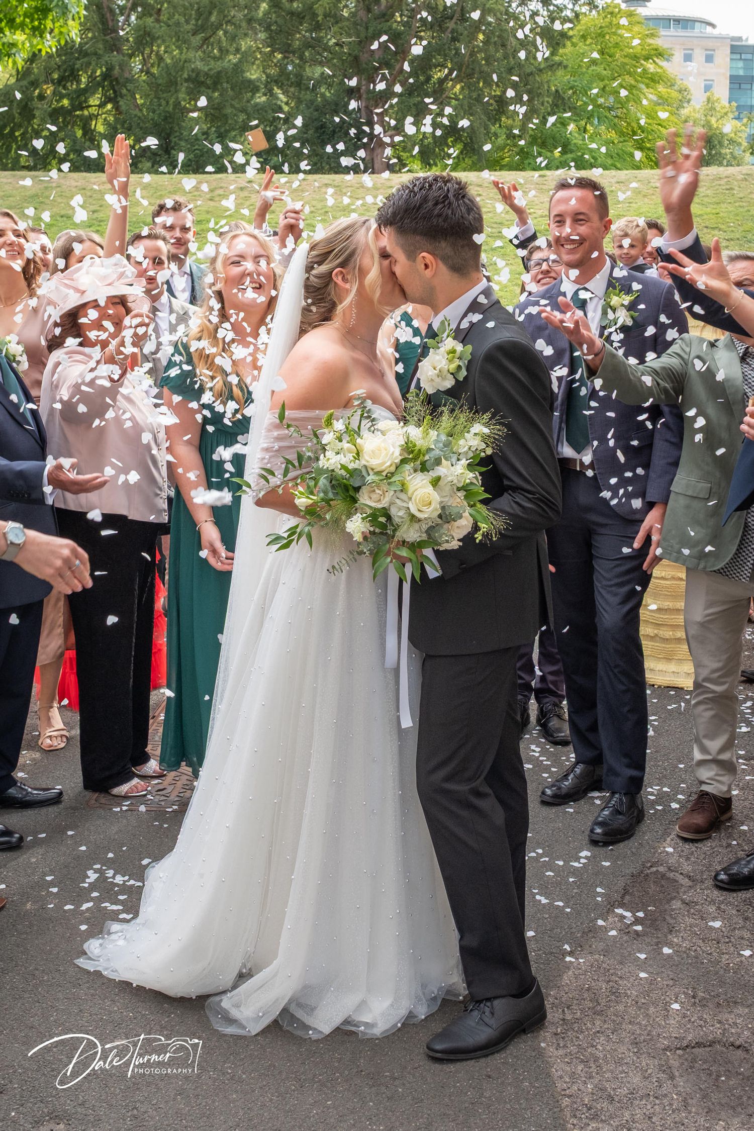 Wedding couple surrounded by guests throwing white confetti outside stone building.
