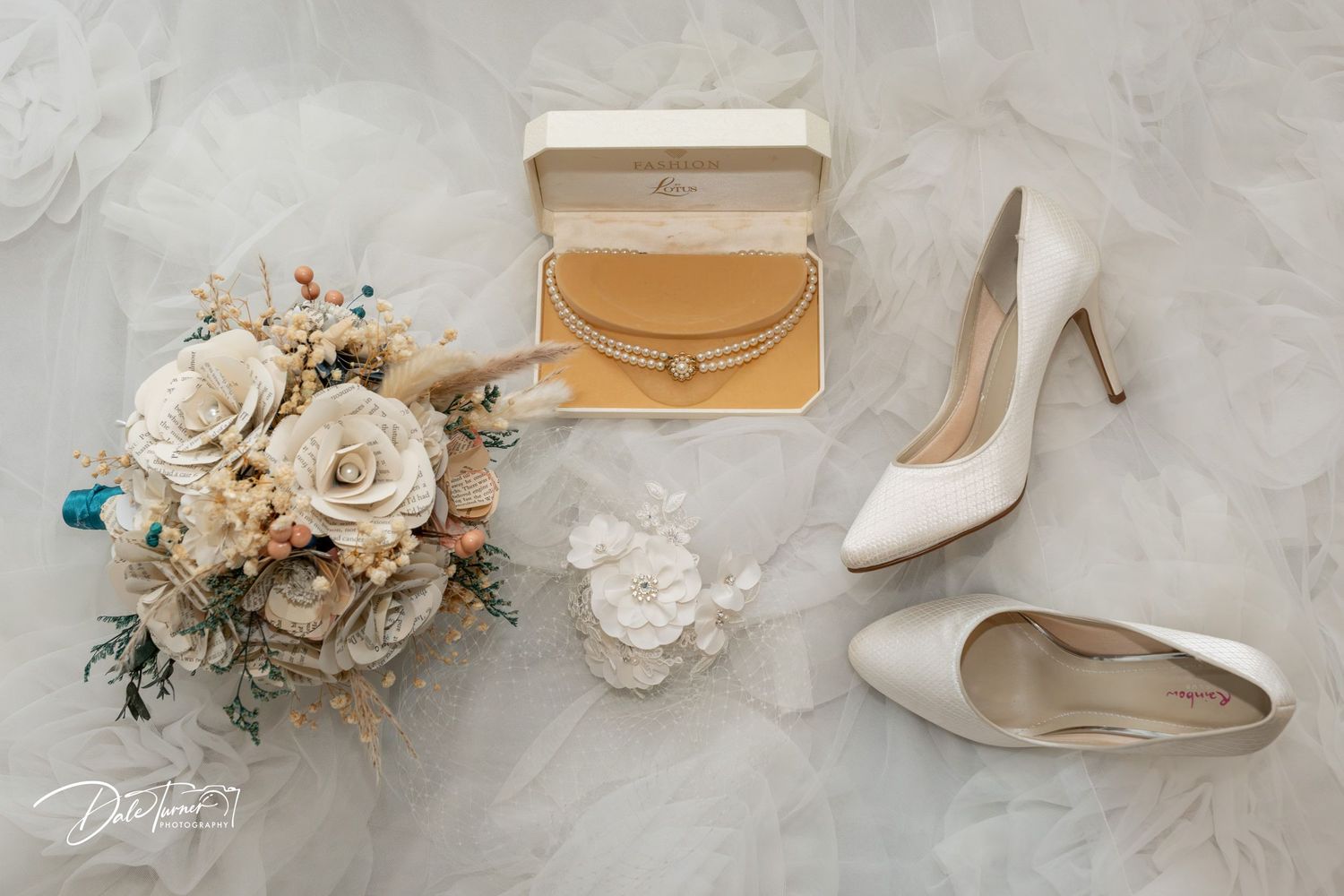 Elegant wedding flat lay featuring white heels, ivory rose bouquet, and gold jewelry box on marble background.
