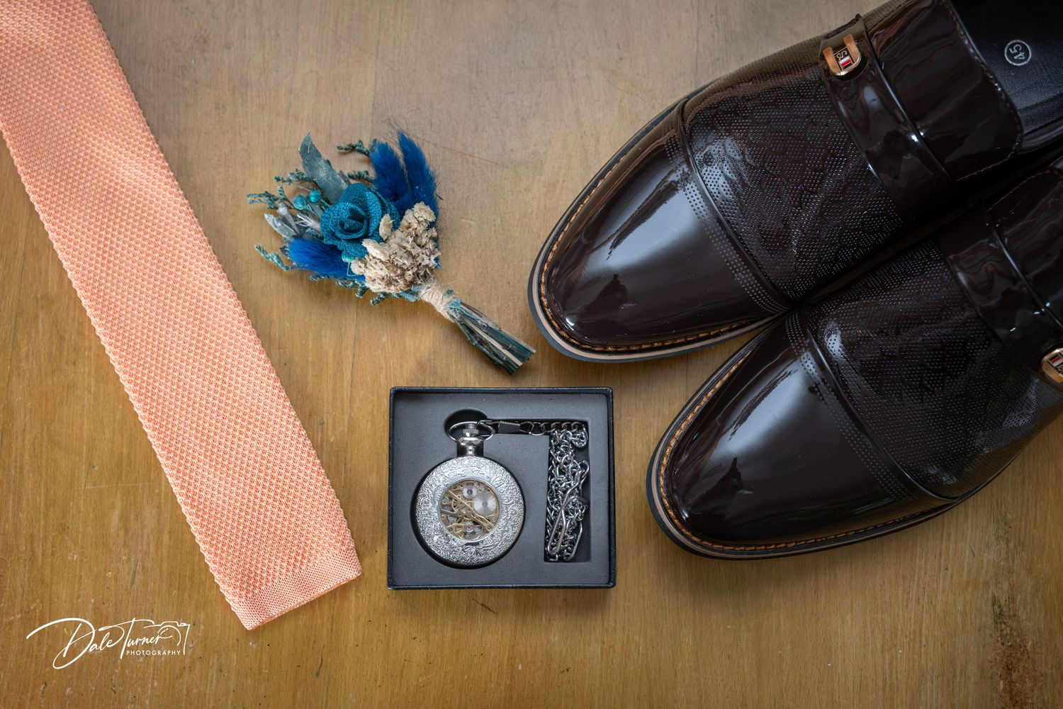 Groom's wedding accessories flat lay featuring brown dress shoes, pocket watch, boutonniere, and salmon knit tie on wood.