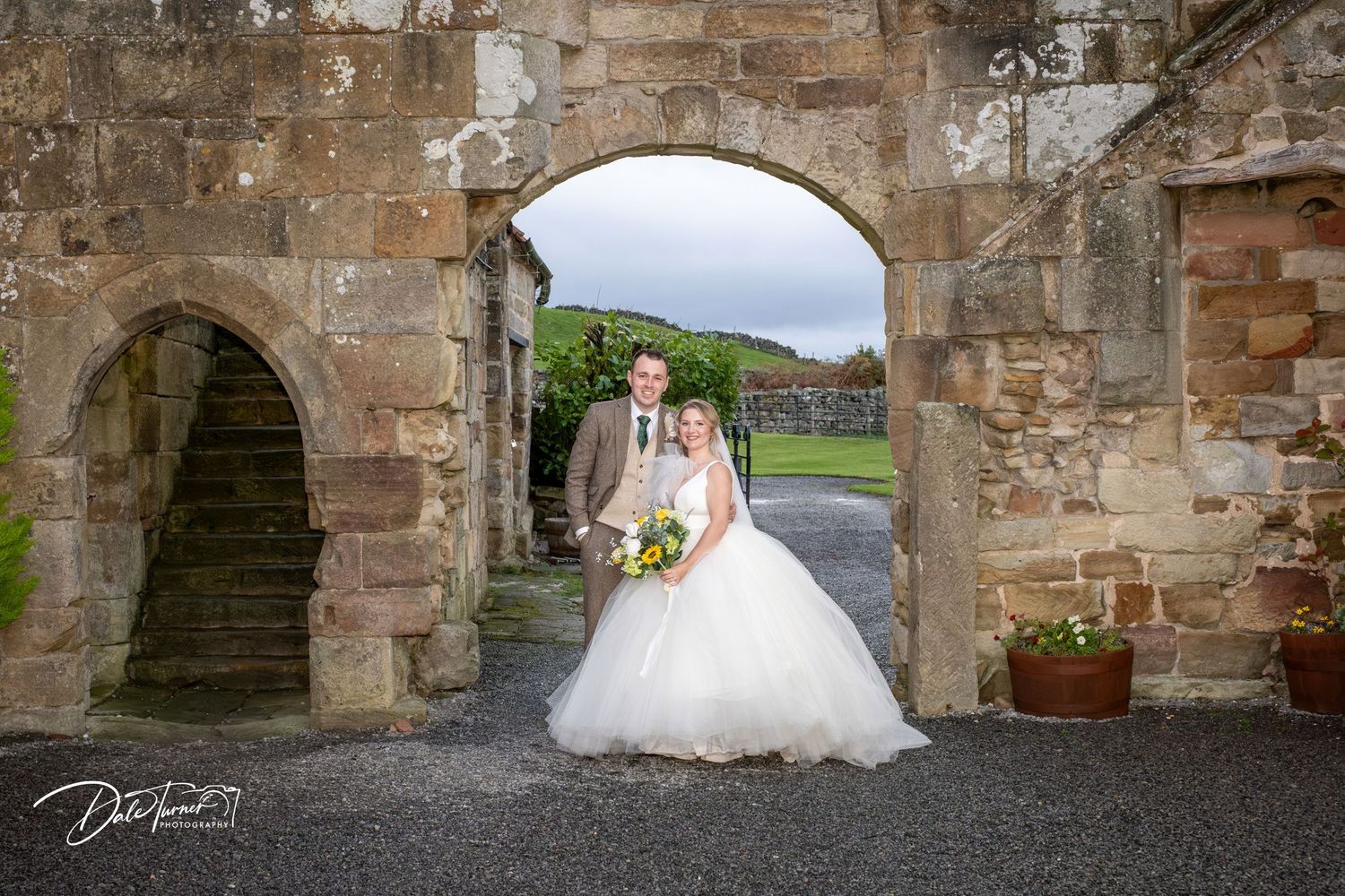 A couple stands beneath a stone archway in a rustic setting with green hills visible in the background.