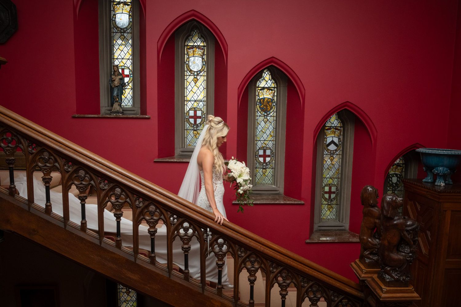 A bride descends a Gothic-style staircase with stained glass windows and rich red walls.