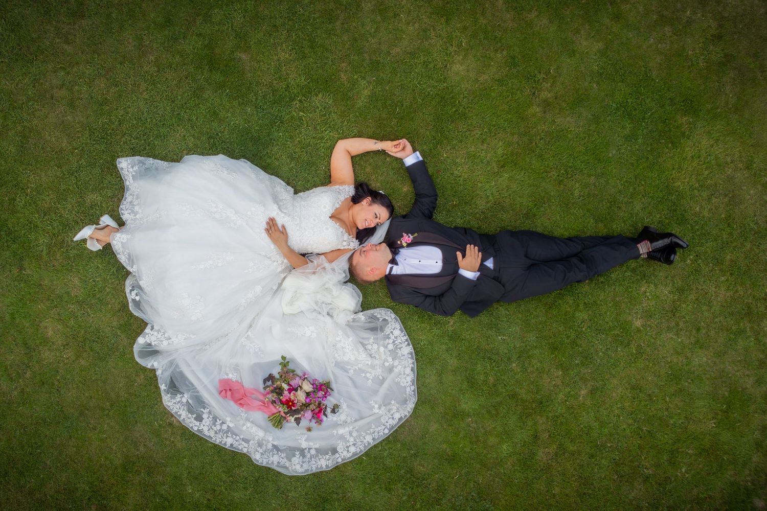 Aerial view of couple lying on green grass with flowing white wedding dress spread out.
