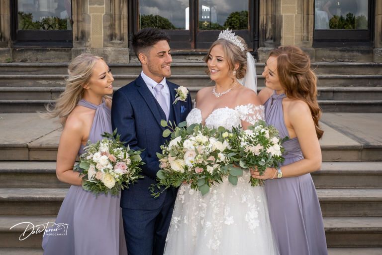Wedding celebration group poses outdoors on stone steps of historic building.