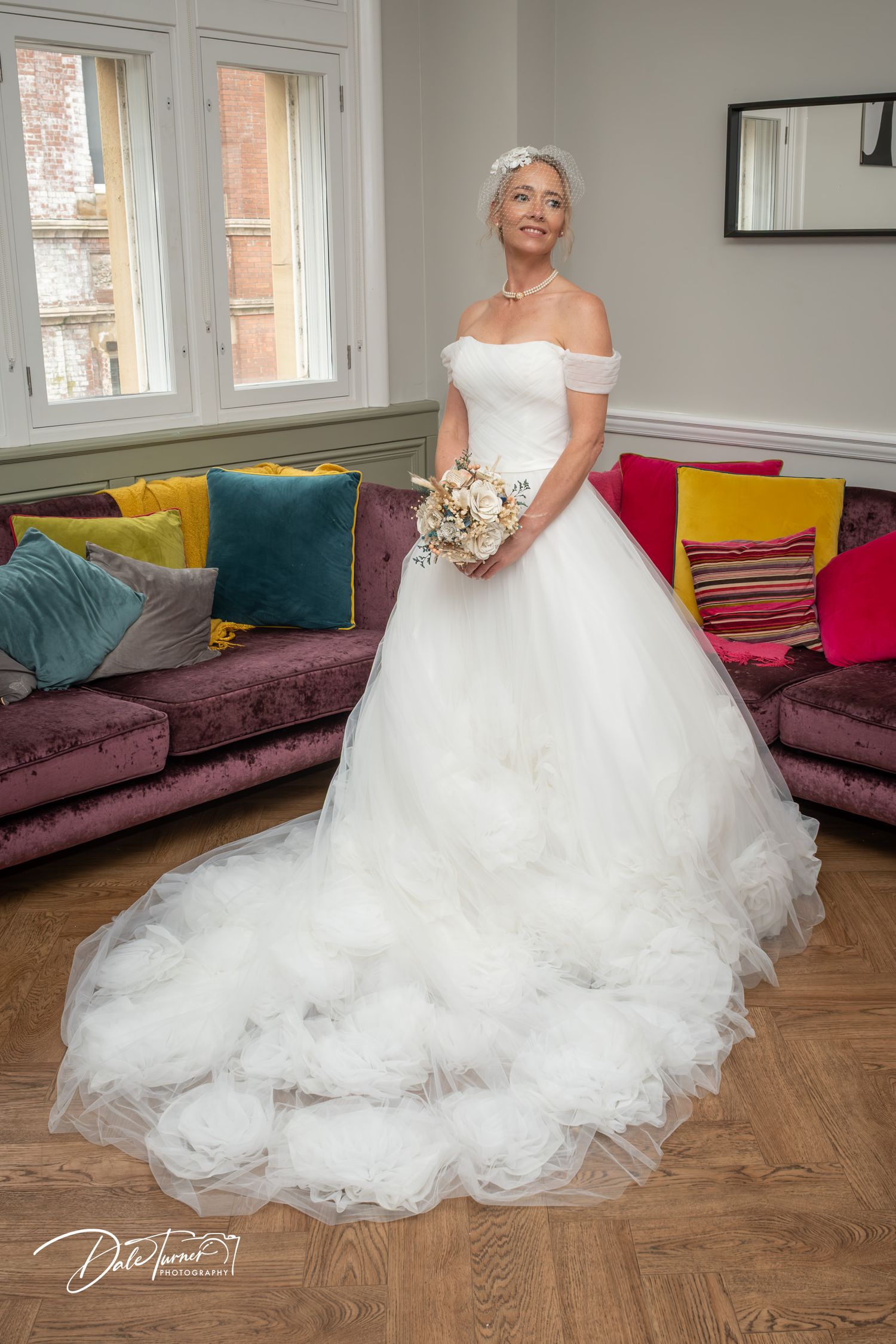 Bride in elegant off-shoulder white ballgown with ruffled train holds bouquet, standing in a colorful room.