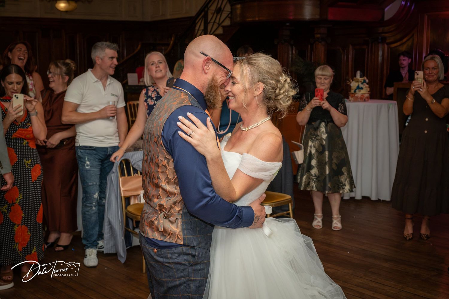 Bride and groom share their first dance surrounded by smiling wedding guests in an elegant venue.