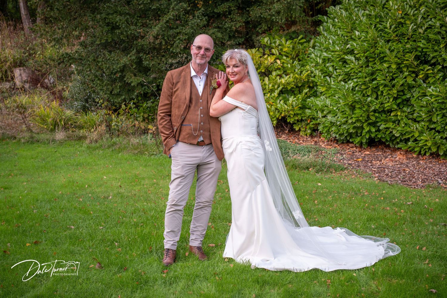 Wedding couple poses on green lawn with autumn foliage in background.