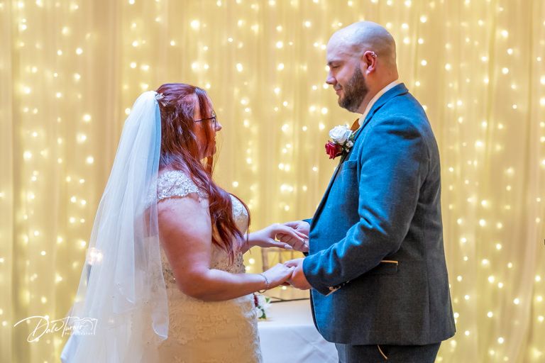 Couple exchanges rings during ceremony in front of romantic fairy light backdrop.