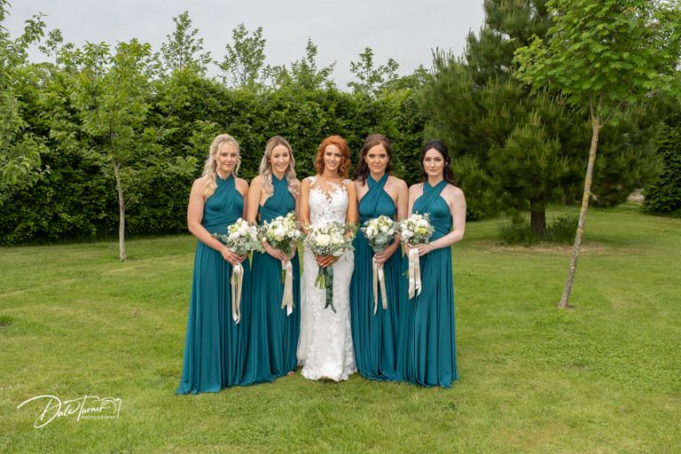 Bridesmaids in teal blue dresses pose together on a lush green lawn during a summer wedding.