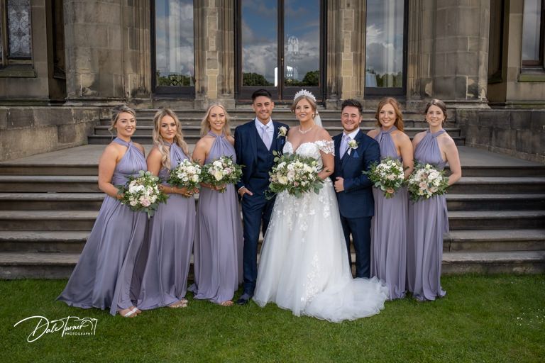 Wedding party in navy and lavender attire gathered on lawn in front of grand stone mansion.