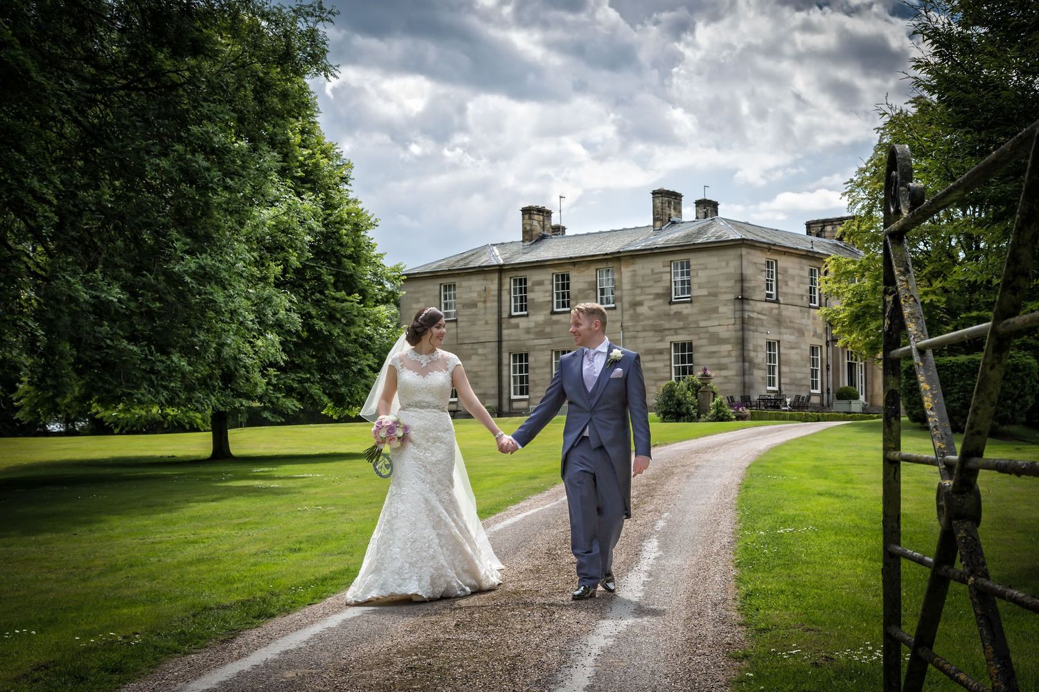 Wedding couple walking along a scenic path with dramatic cloudy sky and historic building backdrop.