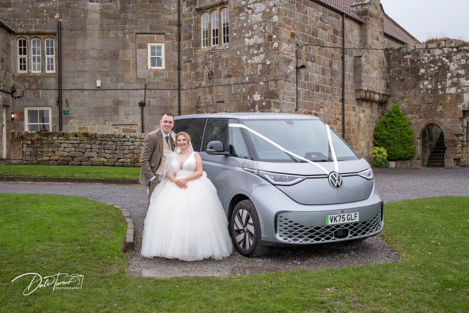 A bride in a white wedding gown poses next to a silver Volkswagen car outside a stone building.
