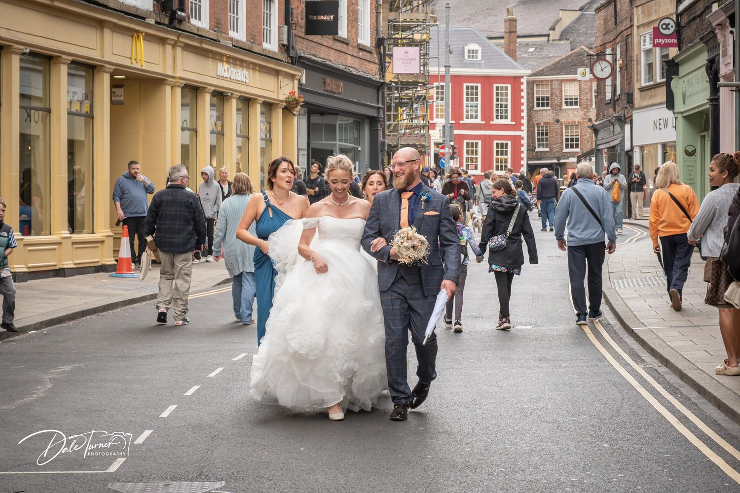 Bride in white gown walks down a busy UK street with companions on her wedding day.