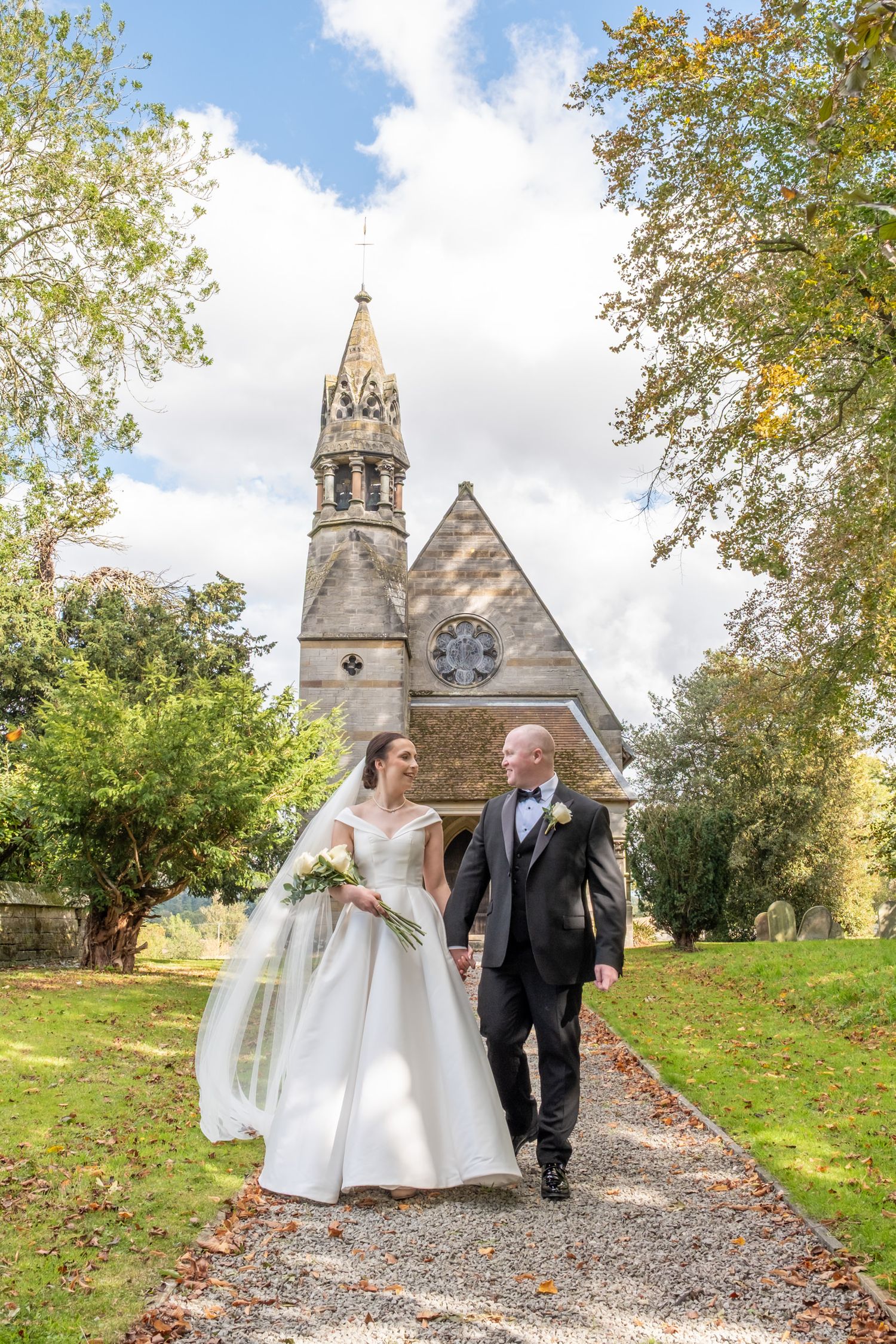 Wedding couple walking along a gravel path towards a historic stone church with autumn trees.
