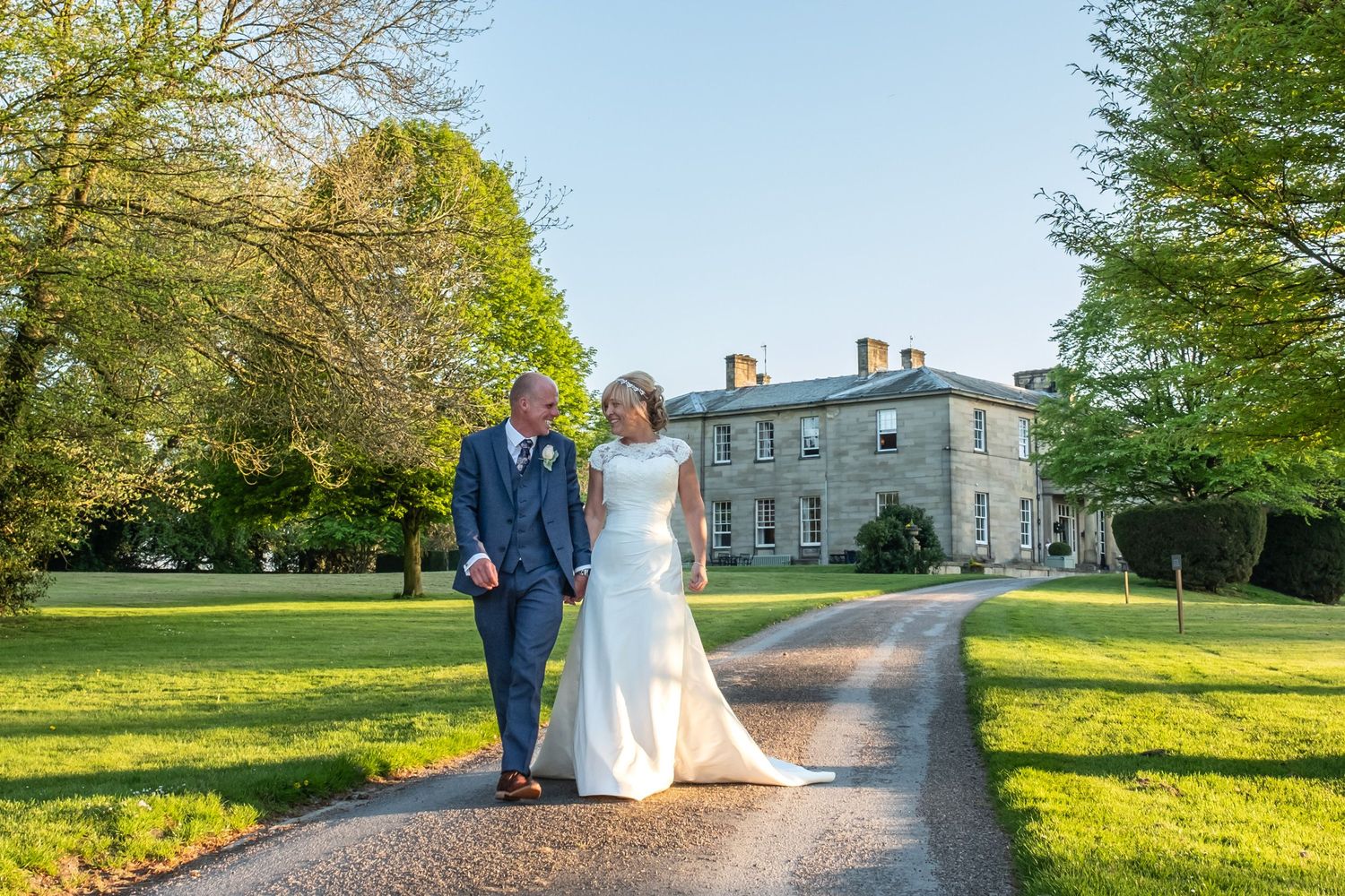 Elegant wedding couple walking along a tree-lined path at a stately manor house on a sunny autumn day.