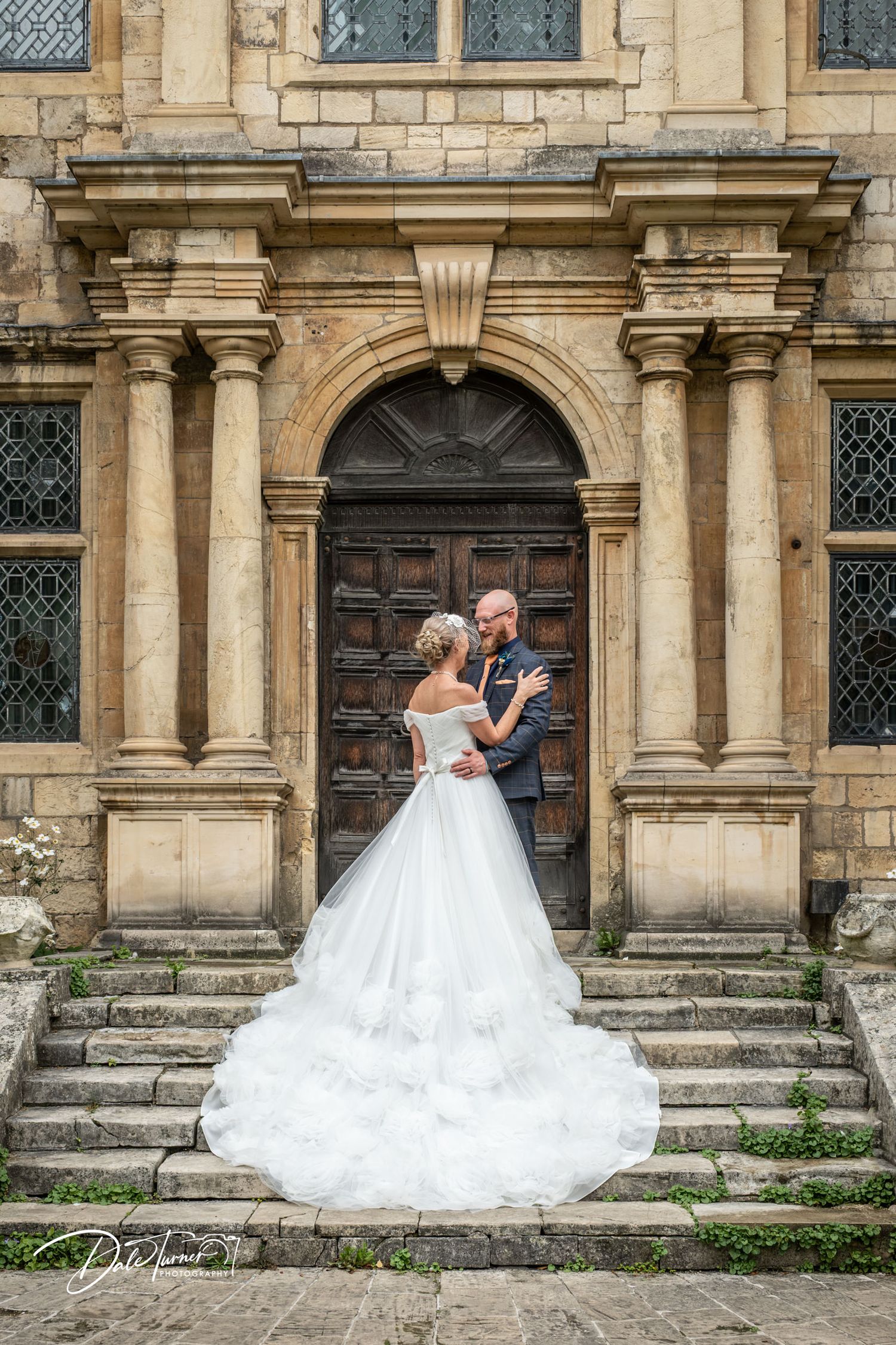 Bride and groom embrace on stone steps before ornate arched doorway of historic venue, wedding dress train spread elegantly.