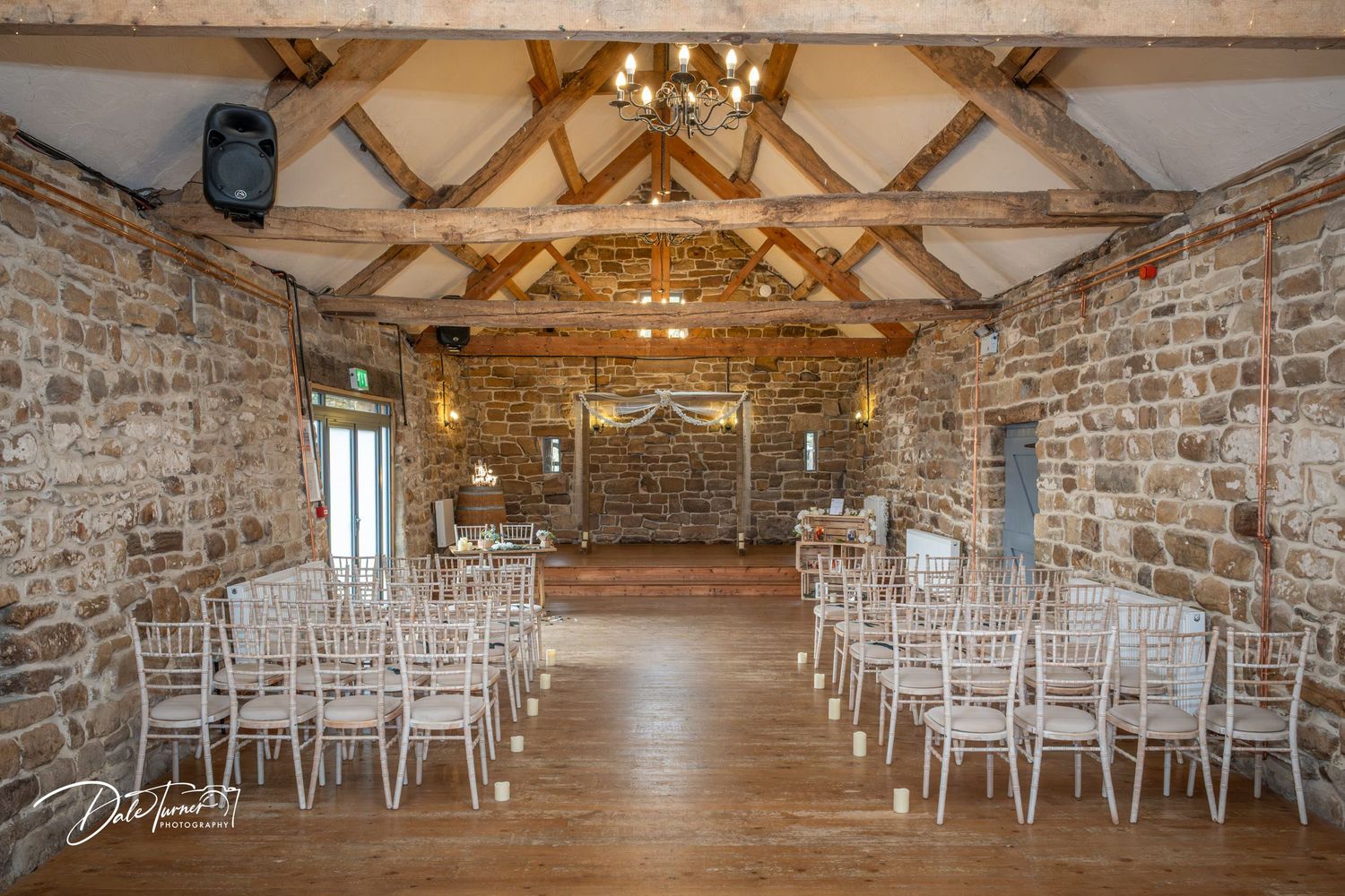 Rustic barn wedding venue interior with exposed stone walls, wooden beams, and rows of white chairs for ceremony setup.
