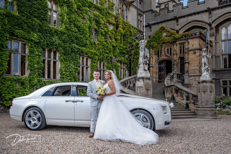 White vintage car parked in front of stone Gothic architecture with ivy-covered walls.