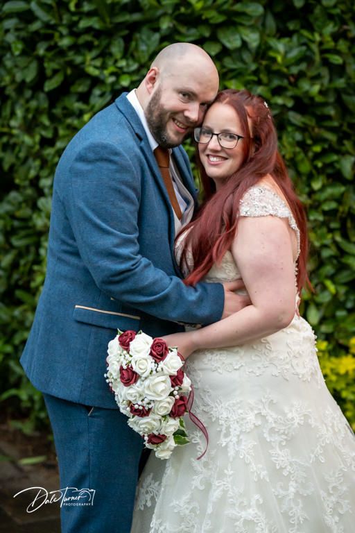 Wedding couple poses in garden setting with white and burgundy floral bouquet.