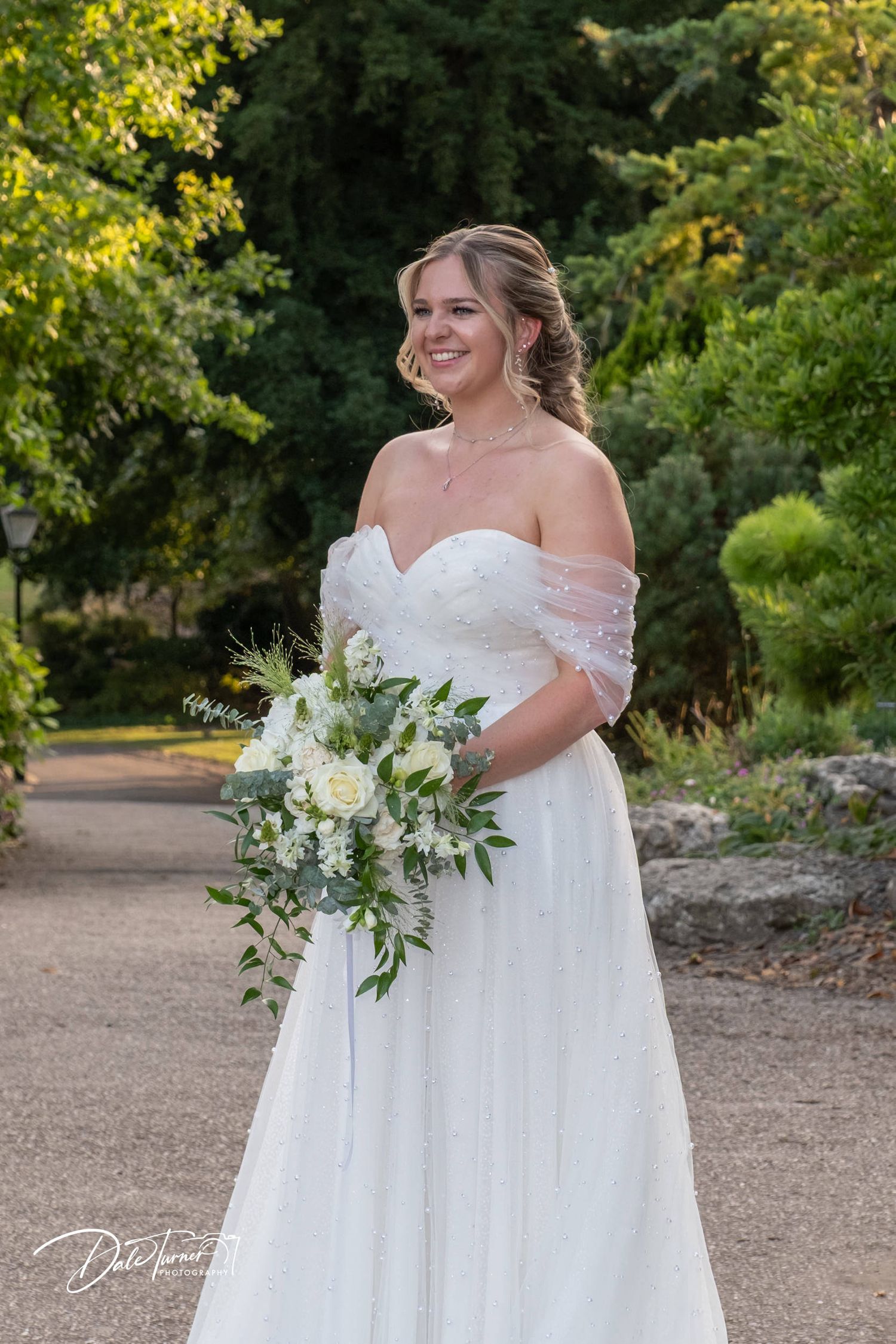 A bride in an off-shoulder gown holds a lush white and green bouquet in a garden setting.
