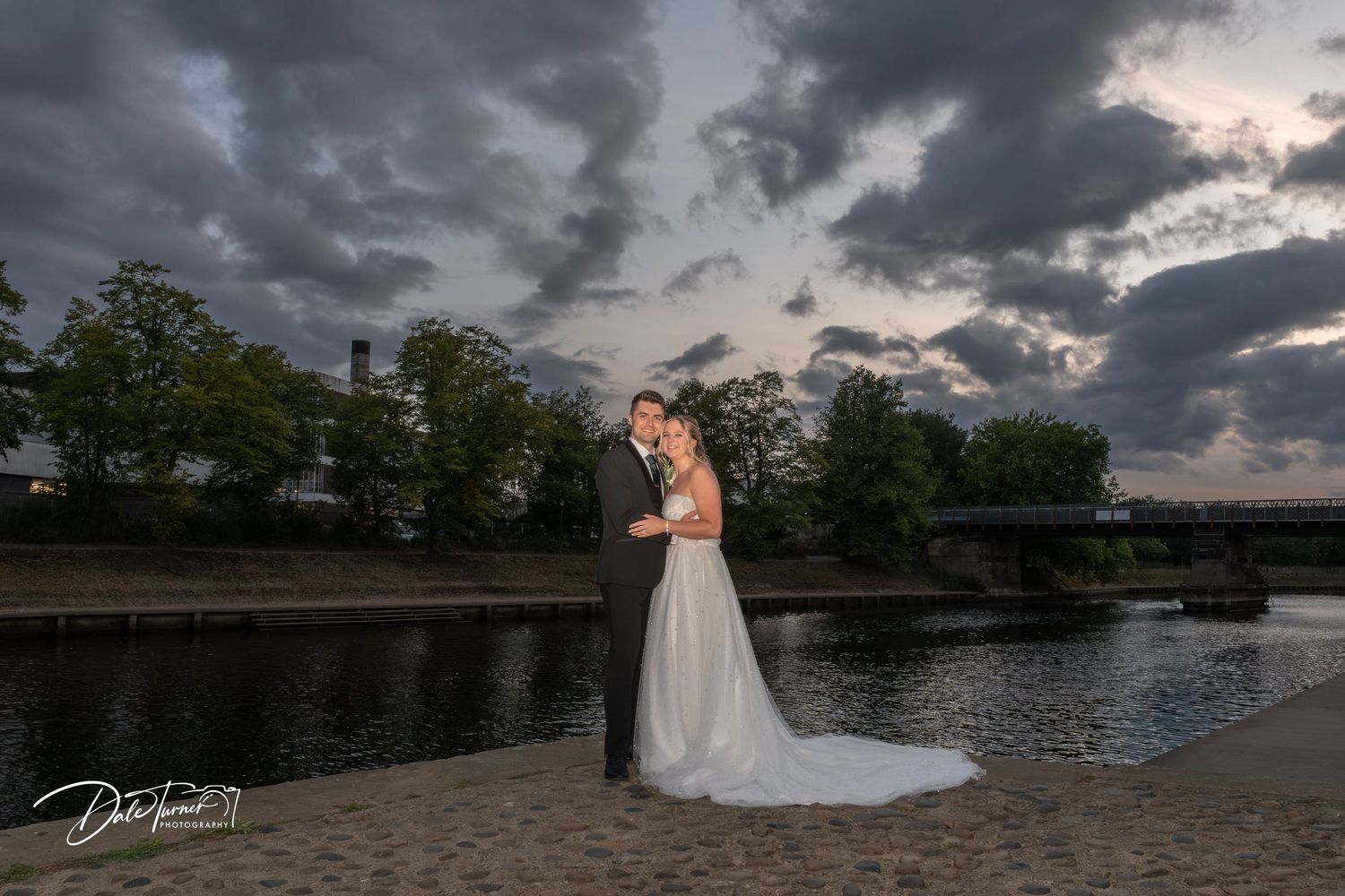 Couple embraces at sunset by a waterfront with dramatic storm clouds in the background.