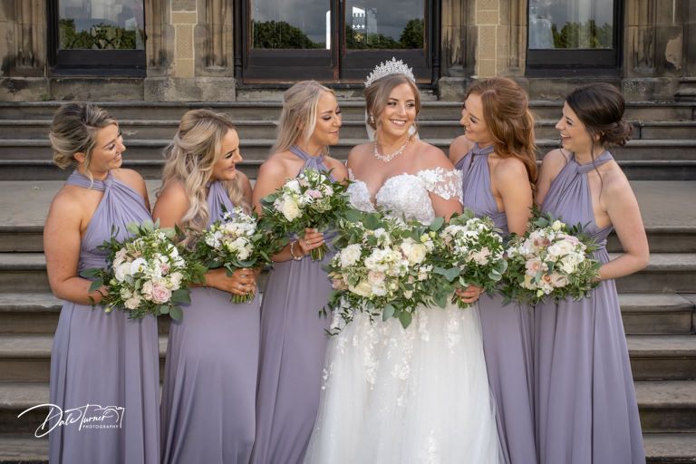 Wedding party in lavender bridesmaid dresses poses on grand stone steps with white floral bouquets.