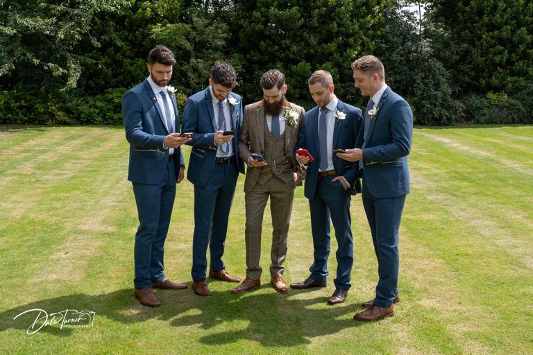 Groomsmen in coordinated navy suits gather for casual photos on green lawn.