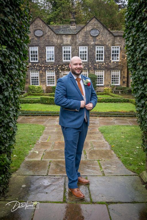 Groomsman in navy suit stands on wet stone patio in front of elegant historic mansion.