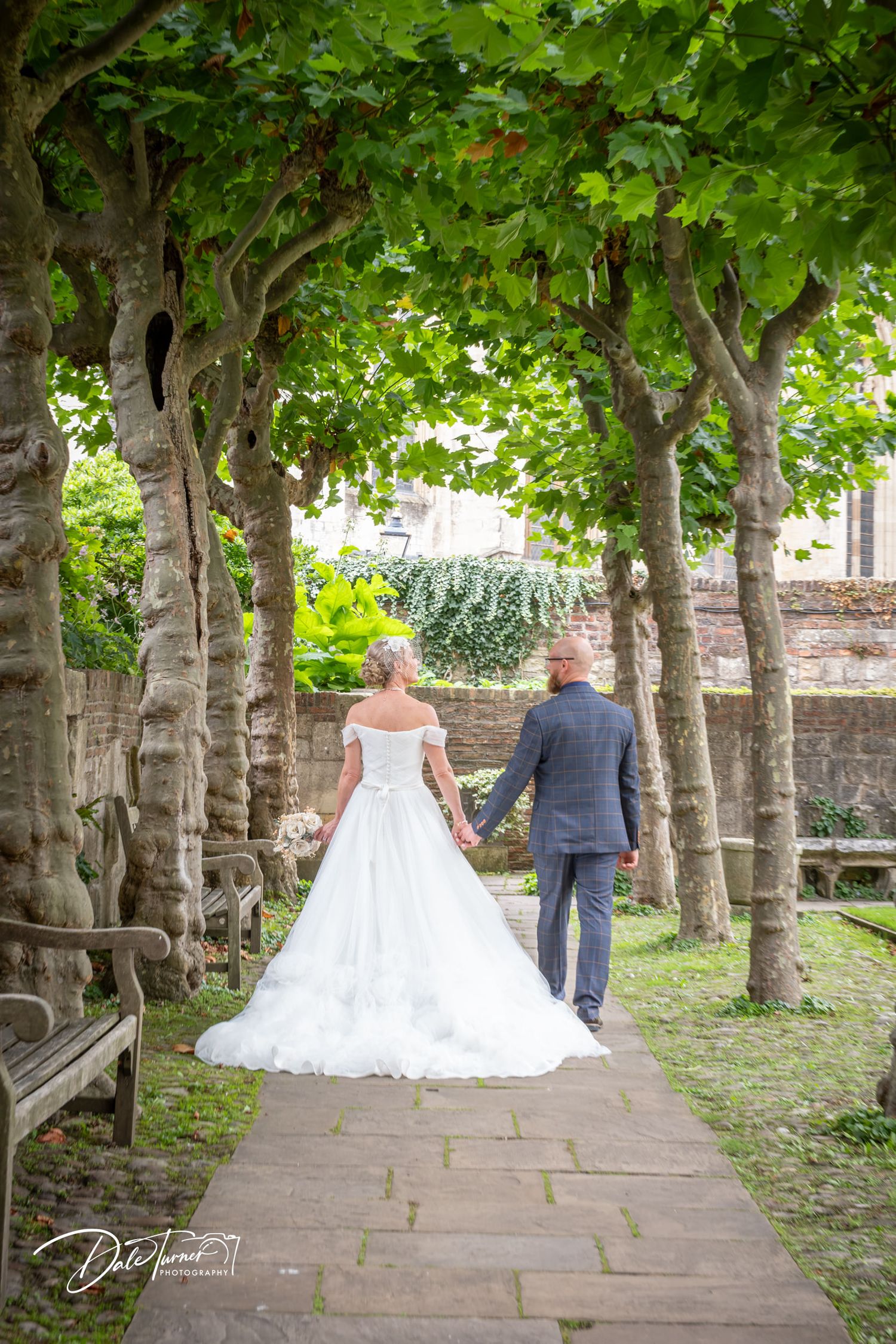 Bride and groom walking hand in hand along a tree-lined garden path, bride in elegant white ball gown.