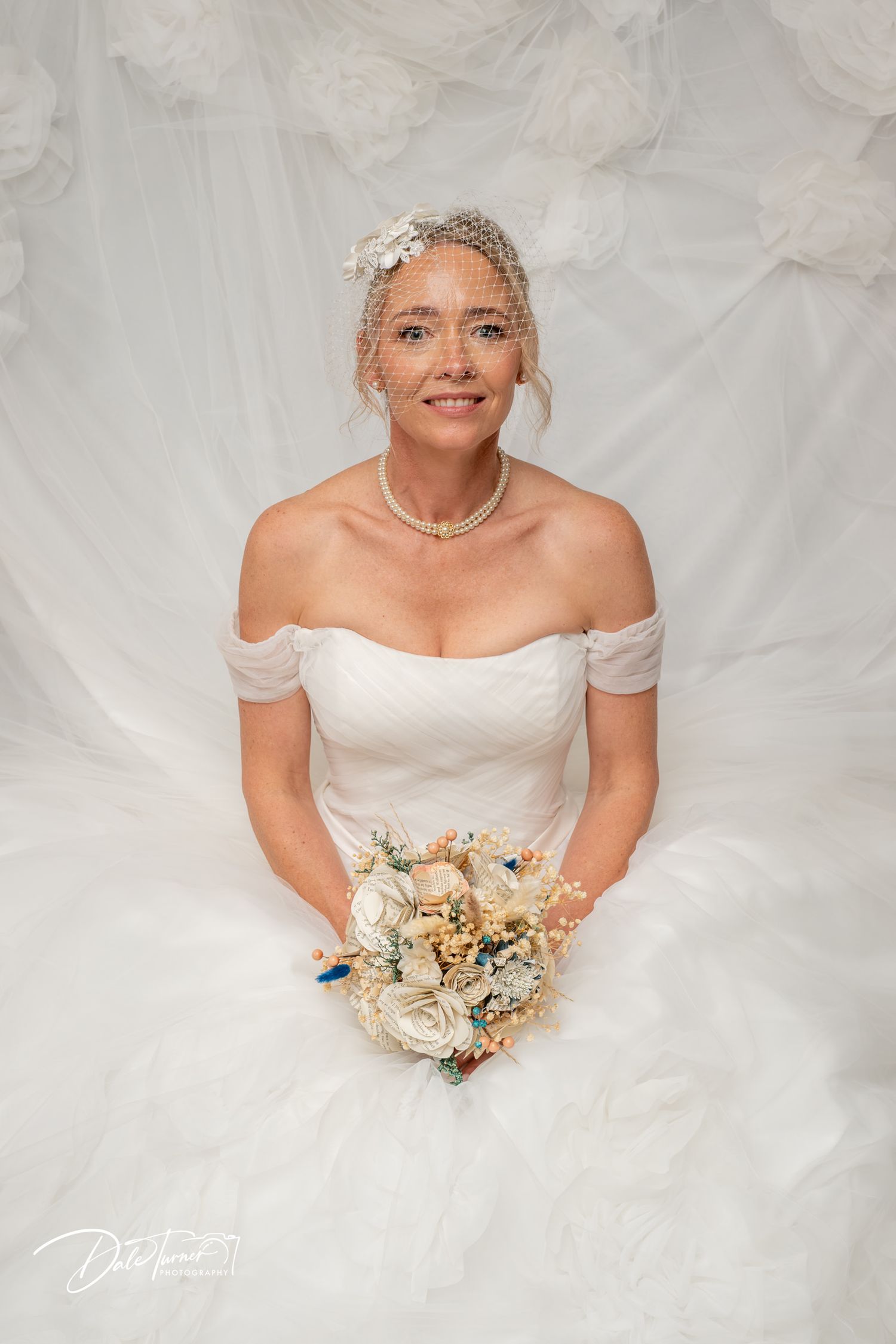 Bride in off-shoulder white gown holds ivory bouquet, wearing birdcage veil, seated against white floral backdrop.